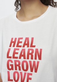 Camiseta de algodón blanca con texto rojo en negrita: "SANAR APRENDER CRECER AMAR". Cuello redondo y mangas cortas, textura suave.