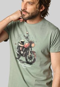 T-shirt en coton vert clair avec un graphique de moto et un casque. Le texte indique "Sunset Expedition Deluxe," imprimé en blanc.