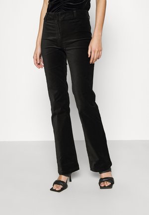 Pantalons en velours côtelé noirs évasés avec une taille haute, texture lisse et subtiles lignes verticales. Portés avec des sandales à talons noires.