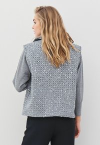 Gray vest with a textured surface, adorned with silver sequins. Features a collar and sleeveless design, paired with a gray long-sleeve top.