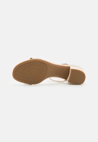 Sandale à talon couleur crème avec une semelle en caoutchouc marron texturée, comportant de fines lanières et un talon bloc. La semelle présente un motif géométrique.