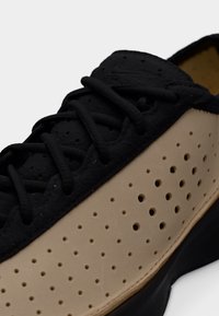 Κοντινό πλάνο μπεζ και μαύρου αθλητικού παπουτσιού με διάτρητο πλαϊνό πάνελ και μαύρα κορδόνια σε μαύρη γλώσσα και σόλα.