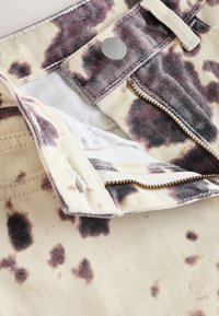 Shorts en denim beige avec un motif éclaboussé violet foncé, fermeture par bouton argenté et fermeture éclair. L'intérieur est doublé en blanc.