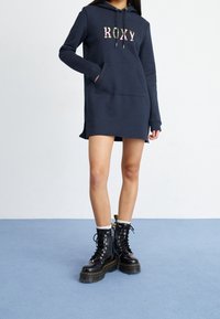 Robe sweat à capuche bleu marine avec un logo "ROXY" floral, manches longues, poche kangourou, fentes latérales, associée à des bottes de combat noires à lacets.