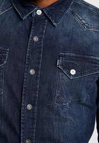 Camicia in denim blu scuro con apertura anteriore a bottoni, colletto a punta, doppie tasche sul petto e bottoni a pressione argentati. Presenta una texture sbiadita.