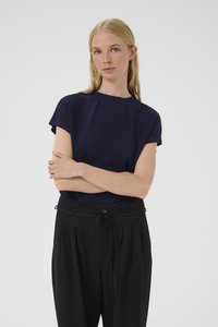 Blondýna s rovnými vlasy má na sobě tmavě modrý krátký rukáv a vysokým pasem černé kalhoty, stojí se založenýma rukama na bílém pozadí.