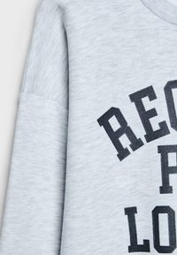 Chemise gris clair à manches longues en gros plan montrant une manche repliée et un texte noir partiel imprimé en lettres majuscules grasses.