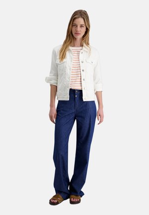 Woman wearing a white denim jacket over a white and orange striped shirt, dark blue wide-leg jeans, and brown sandals, standing against white background.