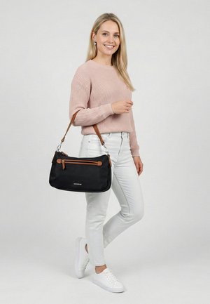 Mujer sonriente con suéter rosa y vaqueros blancos sosteniendo un bolso negro con correa marrón, de pie frente a un fondo claro y liso.