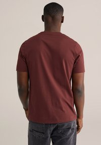 Bordeauxrode T-shirt met korte mouwen, gemaakt van katoen, met een ronde hals en een gladde textuur, gezien van achteren tegen een neutrale achtergrond.