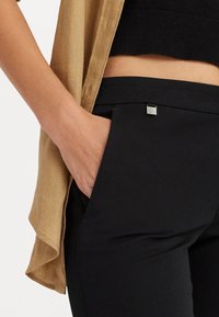 Zwarte strakke broek van een glad materiaal, met een zijzak en een klein metallic label. Gelaagd met een lichte, tan kleurige shirt.