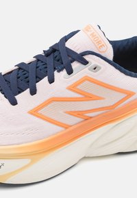 Zapatilla de color morado claro con acentos naranjas, cordones en azul marino y tela texturizada. Presenta una suela blanca gruesa con detalles de patrón esculpido.