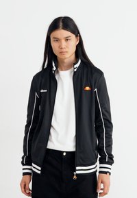 Sort træningsjakke med hvide detaljer, ribbede ærmer og krave. Har et lille rødt og orange logo på brystet. Glat stofstruktur.