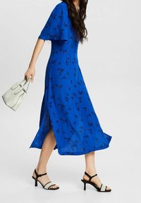 Modrá květinová maxi šaty s krátkými rafinovanými rukávy, bočním rozparkem, doplněné černobílými sandály na podpatku a bílou kabelkou.