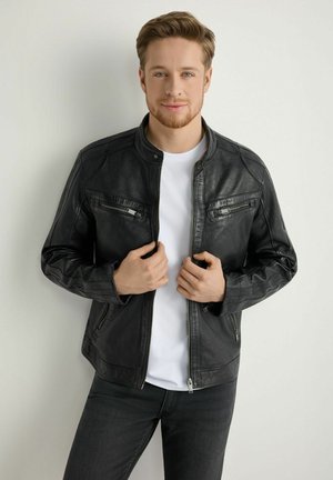 Młody mężczyzna o jasnych włosach, ubrany w czarną skórzaną kurtkę i białą koszulę, stojący na tle jednolitego, jasnego tła.