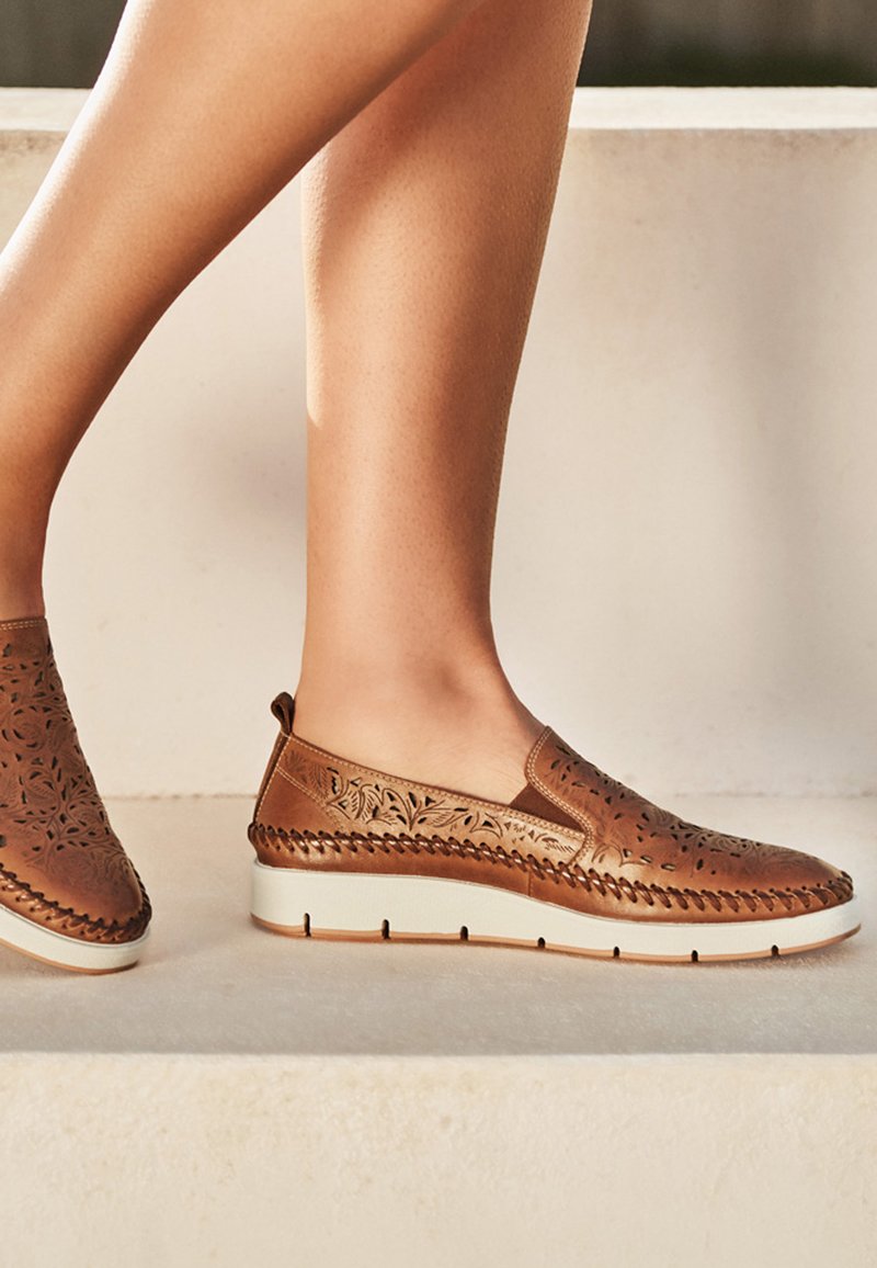 Jambes portant des chaussures en cuir marron sans lacets avec des motifs floraux découpés et des semelles blanches, debout sur des marches en pierre beige.