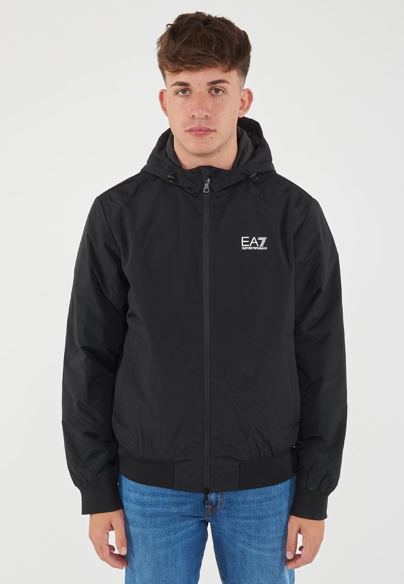 Black hooded jacket with a zip front, elastic cuffs, and a logo on the left chest, paired with blue denim jeans. Smooth texture and modern fit.