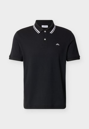 Man draagt een zwart poloshirt met een witte gestreepte kraag, beige wijde broekspijpen, witte sneakers, en houdt een witte handschoen in zijn rechterhand.