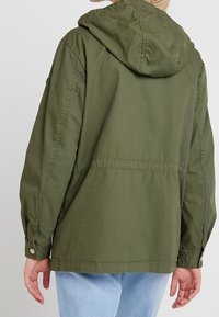 Olive green, oversized jacket with a hood. Features a drawstring waist, buttoned cuffs, and a smooth, lightweight fabric texture.