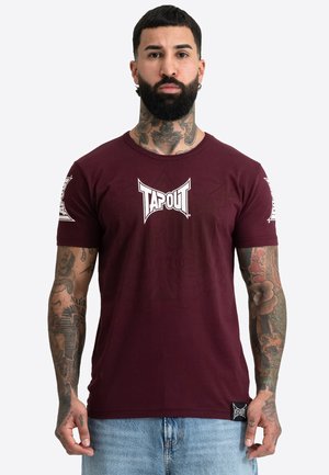 Muž s bradou a tetovaniami na celej ruke, ktorý má na sebe bordové tričko Tapout a svetlomodré rifle, stojí pred jednoduchým pozadím.