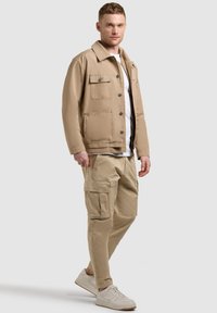 Homme debout portant une veste beige à boutons, un pantalon cargo assorti, un t-shirt blanc et des baskets blanches sur un fond gris uni.