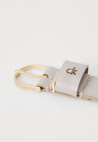 Calvin Klein Gürtel mit einem hellgrauen Lederband, goldfarbener Schnalle, geprägtem Logo und Stichdetails. Durchgehend glatte Textur.