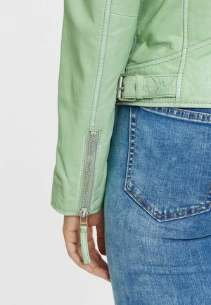 Grüne Lederjacke mit tailliertem Design, seitlichem Reißverschluss und Schnallenakzent. Getragen über blauen Jeans mit Gesäßtasche.