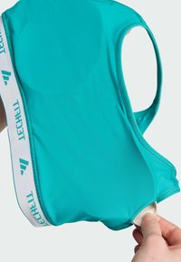 Tirkizni sportski grudnjak od mekog materijala, s racerback dizajnom i bijelom elastičnom trakom s plavim akcentima. Glatka textura s bešavnim rubovima.