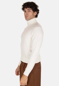Maglione a collo alto bianco a coste con un design aderente, dotato di un alto colletto e maniche lunghe, indossato con pantaloni marroni.