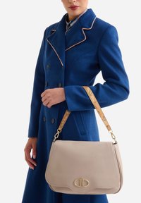 Borsa a mano in pelle beige con una forma curva, ferrature dorate e una tracolla decorata. Indossata con un cappotto blu con rifiniture e bottoni a contrasto.