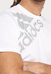 Camiseta atlética blanca con un patrón gráfico texturizado negro en la zona del pecho y los hombros, con escote redondo y mangas cortas.
