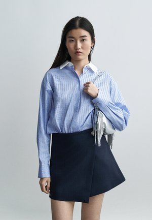 Νεαρή γυναίκα που φοράει ένα φαρδύ μπλε ριγέ πουκάμισο, μαύρη ασύμμετρη φούστα και κρατά μια ασημένια τσάντα με φούντες στον ώμο της.