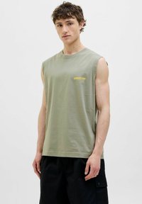 Olivenfarvet ærmeløs t-shirt i blød bomuld med rund hals; har et gult trykt logo på venstre bryst. Sorte cargo-bukser er synlige.
