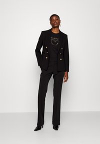 Blazer nero su misura con chiusura doppiopetto e bottoni dorati, abbinato a pantaloni neri coordinati e a un top nero aderente con decorazioni.
