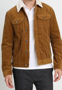 Brun kordfløjljakke med en crèmefarvet fårskindsfarvet krave, som har to brystlommer og knaplukning, tekstureret stof med ribbede mønstre.