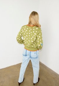 Γυναίκα με μακριά ξανθά μαλλιά, που φοράει ένα πράσινο πουλόβερ με πουά και ανοιχτό μπλε τζιν, στέκεται με τα χέρια στη μέση, κοιτάζοντας έναν λευκό τοίχο.