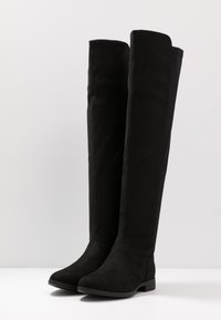 Bottes noires hautes en suède doux, avec une semelle plate et un bout rond, présentant des lignes épurées et un design minimaliste.