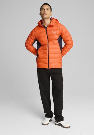 Orange pufferjakke med hætte, der har mørkeblå sidelapper. Lynlåslukning og et lille logo på brystet. Båret over sorte bukser.