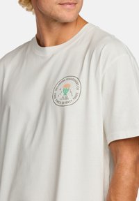 Camiseta beige claro de algodón suave, con un parche de logo redondo que presenta diseños en verde y naranja en el área del lado izquierdo del pecho. Mangas cortas.