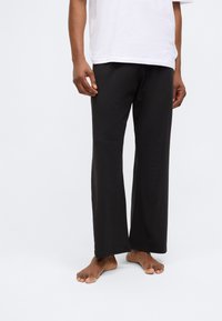 Pantalons larges noirs en tissu léger, présentant une coupe droite et une finition lisse. Portés avec un t-shirt blanc ample.