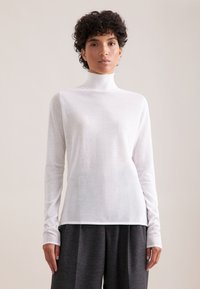 Biała, długosleeve'owa bluzka z golfem z lekkiego materiału, o dopasowanym kroju i prążkowanym kołnierzem, w zestawieniu z szarymi, luźnymi spodniami.