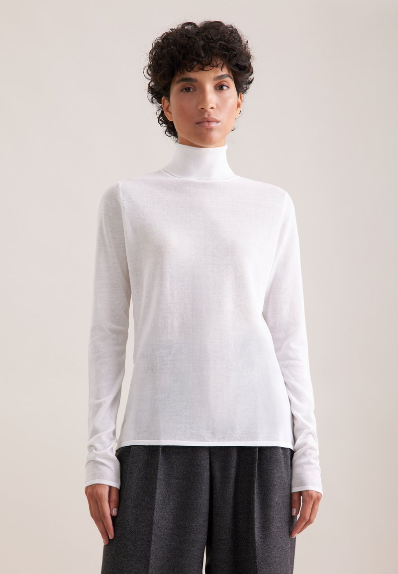 Biała, długosleeve'owa bluzka z golfem z lekkiego materiału, o dopasowanym kroju i prążkowanym kołnierzem, w zestawieniu z szarymi, luźnymi spodniami.