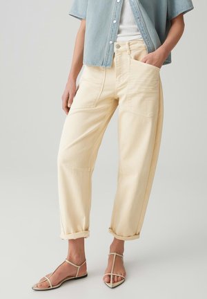 Person trägt hellbeige High-Waist-Hosen mit großen Vordertaschen, aufgekrempelten Bündchen, kombiniert mit beigen, flachen Sandalen mit Riemchen.