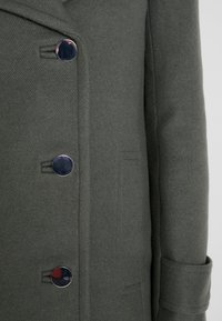 Abrigo de lana verde oscuro con una tela texturizada, que presenta botones plateados brillantes y un bolsillo lateral. Diseño clásico de solapa con muesca.