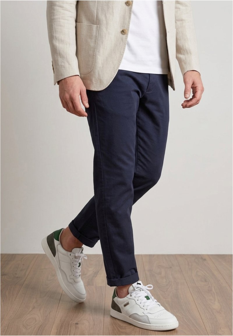 Man draagt een beige blazer, wit T-shirt, marineblauwe omgeslagen broek en witte sneakers met groene en grijze accenten, lopend op een houten vloer.
