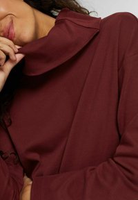 Burgundy pitkähihaisessa paidassa on korkea taitettavakolari ja pehmeä tekstuuri. Siinä on rentoistuvat hihat ja sileä, kevyt kangas.