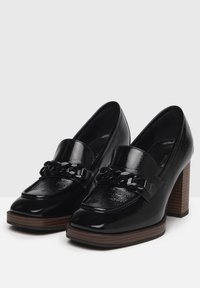 Tacones de charol negro con un chunky bloque de madera, con un acento de cadena en el empeine y un acabado suave y brillante.