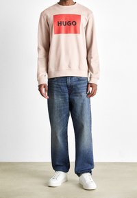 Sudadera rosa claro con un parche rectangular rojo audaz que presenta "HUGO" en negro. Combinada con unos vaqueros azules desgastados y zapatillas blancas.