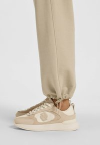 Beige sneakers med ovandel i mocka och textil, rund tå, snörning framtill, dämpad sula och logotyp på sidan. Joggingbyxor som samlas vid vaden.