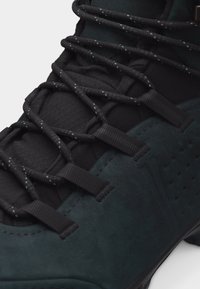 Κοντινό πλάνο από σκούρο πράσινο και μαύρο αθλητικό παπούτσι που δείχνει τη υφή της γλώσσας και τα σφιχτά δεμένα μαύρα κορδόνια με αντανακλαστικά σημεία.